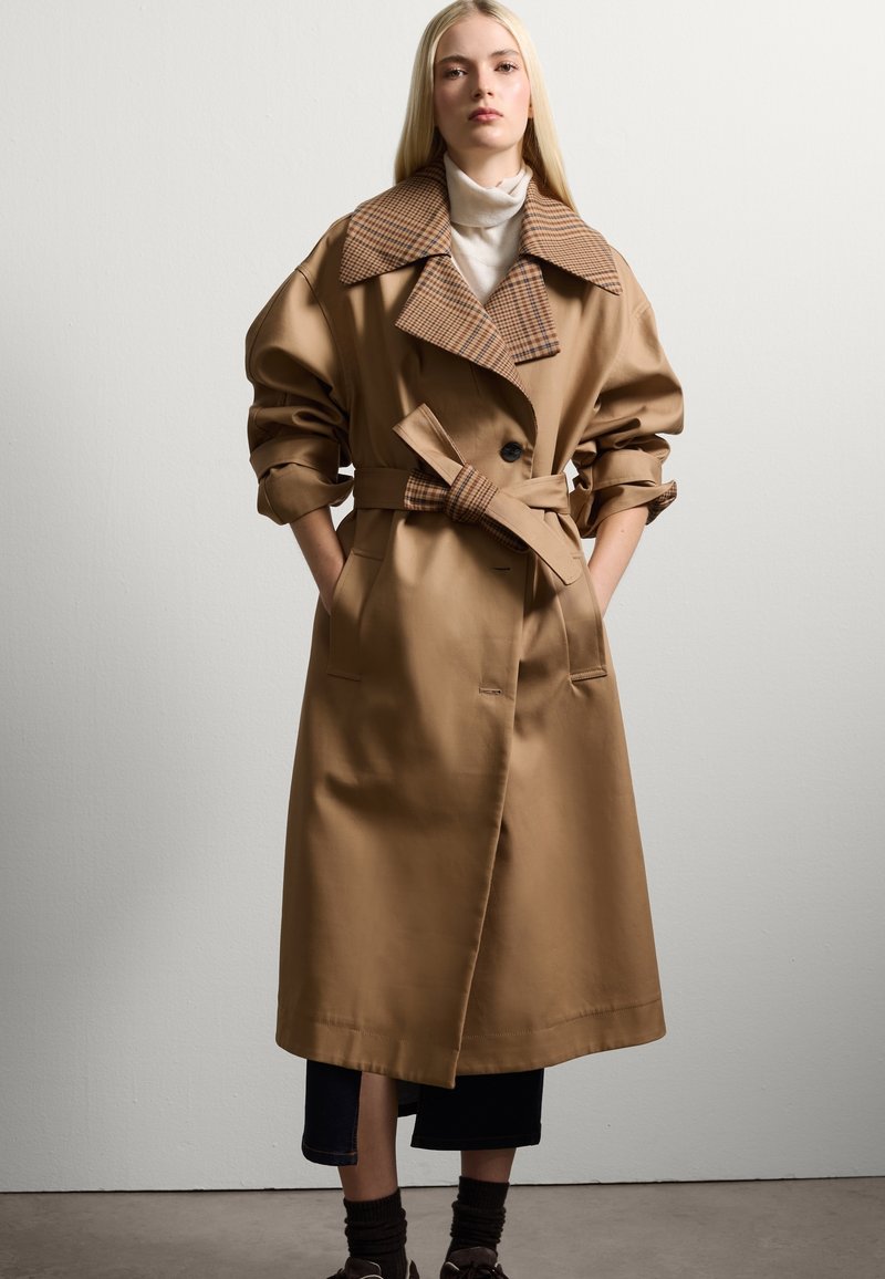 Abrigo trench marrón con un gran cuello de cuadros, mangas anchas, un cinturón atado y mínimos botones. Lleva puesto sobre unos jeans oscuros y zapatos marrones.