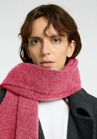 Rosa stickad scarf i mjukt tyg, med ett strukturerat mönster, virad runt halsen och kombinerad med en mörk jacka.