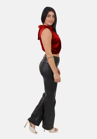 Donna con lunghi capelli scuri, che indossa un top rosso senza maniche, pantaloni in pelle nera, sandali bianchi con tacco alto e un tatuaggio sull'avambraccio.