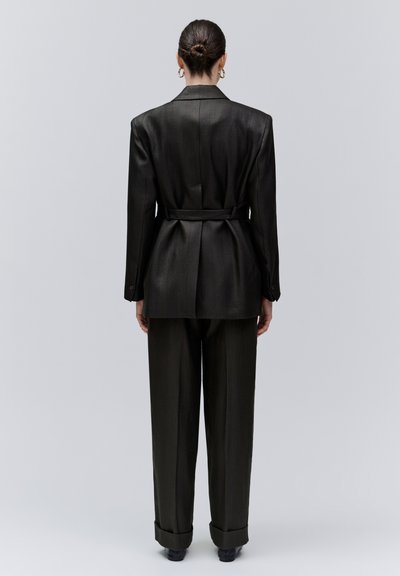 Femme aux cheveux en chignon portant un blazer noir ceinturé et un pantalon large, boucles d'oreilles créoles dorées, debout de dos devant un fond gris uni.