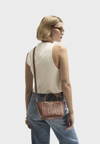 Bruine leren handtas met een geweven textuur, korte handvatten en een afneembare schouderband. Gecombineerd met een lichtgekleurde top en denim jeans.