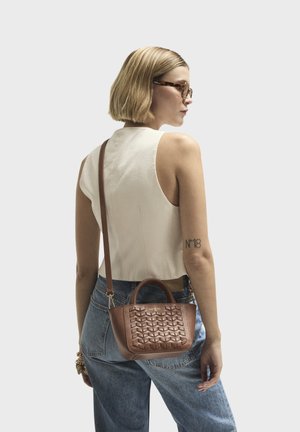 Braune Lederhandtasche mit gewebter Textur, kurzen Griffen und einem abnehmbaren Trageriemen. Kombiniert mit einem hellen Oberteil und Jeans.