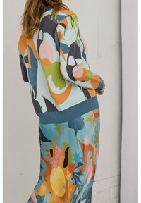 Wielokolorowy kardigan z dzianiny z abstrakcyjnymi wzorami w kolorach zielonym, pomarańczowym, różowym i niebieskim, zestawiony z długą, intensywnie wzorzystą spódnicą z motywem słońca.