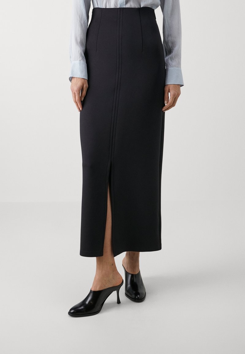 Svart, figurnära maxikjol med en frontslits och subtila sömdetaljer, ihop med spetsiga mules, mot en enkel bakgrund.