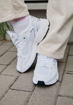 Baskets blanches Adidas portées avec des chaussettes blanches Adidas et un pantalon en velours côtelé beige clair sur un sol pavé près de la verdure.