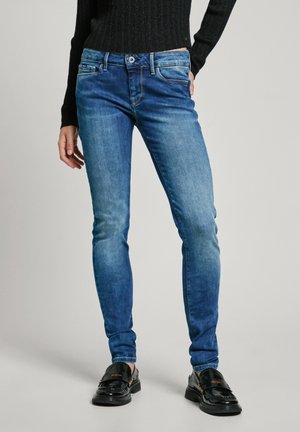 Vrouw in een blauwe slim-fit jeans, zwarte geribbelde trui en zwarte glanzende loafers, staande tegen een effen achtergrond.