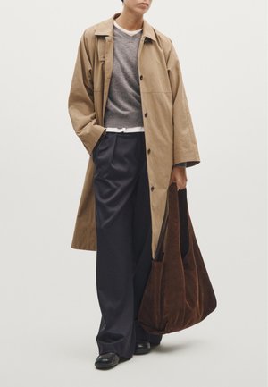 Trench coat beige sopra un maglione grigio, pantaloni larghi blu navy, e tiene una borsa a tracolla in suede marrone. Scarpe nere completano l'outfit.