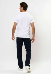 Egy férfi háttal áll, sima fehér pólót, sötét farmernadrágot és fehér sportcipőt visel, amely kék és piros csíkos, egy fehér háttér előtt.