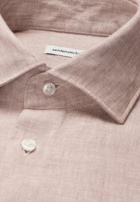 Jasnoróżowa lniana koszula z klasycznym kołnierzykiem i dwoma widocznymi białymi guzikami, starannie złożona z metką marki wewnątrz kołnierzyka.