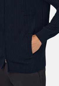 Mornarsko modra rebrasta jopica z zadrgo, stranskimi žepi in teksturnim pletenjem. Narejena iz mehkega materiala z oprijetim krojem.