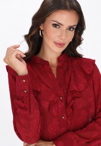 Blusa roja con textura floral, cuello con volantes, cierre de botones en la parte delantera y mangas largas. Detalles en tela transparente y botones plateados.