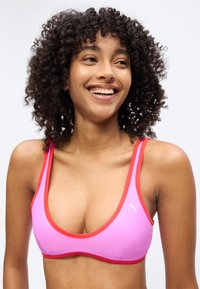 Mosolygó nő göndör hajjal, rózsaszín sportmelltartót visel vörös szegéllyel, kissé oldalra néz egy egyszerű háttér előtt.