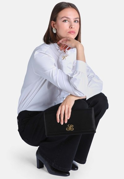 Jeune femme en blouse blanche et pantalon noir accroupie, tenant une pochette noire avec un logo doré, portant de grandes boucles d'oreilles argentées et des chaussures à talons noires.