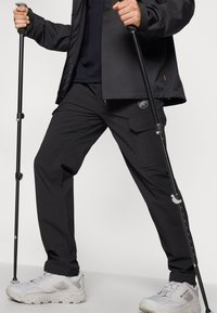 Svarta utomhusbyxor med cargofickor, lättviktsmaterial och elastisk midja. I kombination med vita vandringsskor och vandringsstavar.