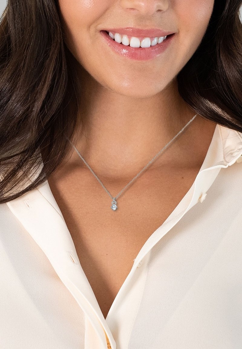 Collier en chaîne en argent mettant en avant un pendentif en pierre précieuse bleue ronde, présenté sur un chemisier de couleur claire.