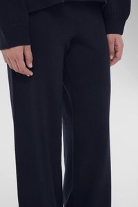 Χέρια ακουμπισμένα στα πλευρά, φοράει σκούρο μπλε μάλλινο παντελόνι και ταιριαστό σκούρο μπλε μάλλινο πουλόβερ πάνω σε απλό ανοιχτό φόντο.