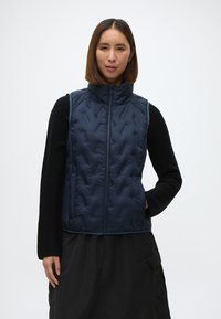 Ναυτικό γιλέκο με πλισέ υψηλό γιακά, κλείσιμο με φερμουάρ και πλαϊνές τσέπες. Φορεμένο πάνω από μαύρο μπλουζάκι με μακριά μανίκια, που διαθέτει υφή.