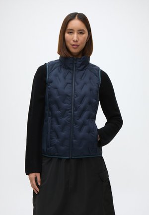 Námornícky prešívaný vest s vysokým golierom, zipsom a bočnými vreckami. Nosený cez čierne dlhé rukávy tričko, so štruktúrovanou tkaninou.