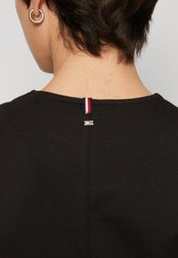 Czarna koszulka z okrągłym dekoltem, z tylnym zapięciem z czerwono-białoniebieską wstążką i małym srebrnym detalem z logo.