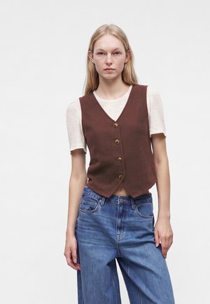 Jeune femme aux longs cheveux blonds portant un gilet marron boutonné par-dessus un haut blanc à manches courtes et un jean bleu sur un fond uni.