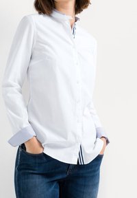 Bílá košile na knoflíky s stojáčkovým límcem, modro-bílé pruhované akcenty a vzorované manžety. Nosí se s tmavě modrými džíny.