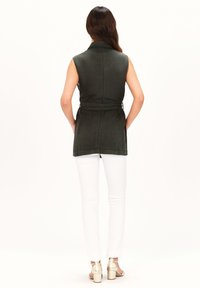 Mouwloos donkergroen vest met een kraag, vastgebonden in de taille. Witte broek en gouden hakken maken de outfit compleet. Gladde textuur, eenvoudig ontwerp.