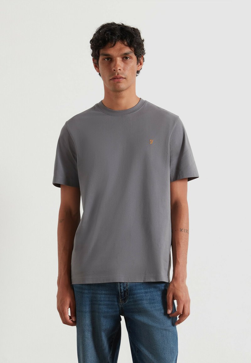 Man met krullend donker haar, gekleed in een eenvoudig grijs T-shirt met korte mouwen en een klein oranje logo op de borst, gecombineerd met een blauwe spijkerbroek.