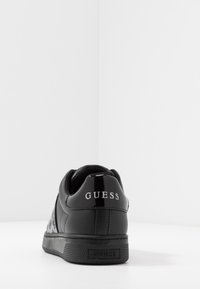Černé nízké tenisky s lesklým zadním poutkem a značením "GUESS" na patě a podešvi, vystavené na bílé ploše na bílém pozadí.