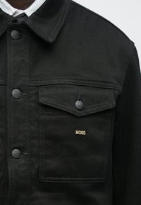Čierna rifľová bunda s dvoma náprsnými vreckami, striebornými kovaniami a malým zlatým logom "BOSS" na ľavom vrecku. Textúrovaná látka s golierovým dizajnom.