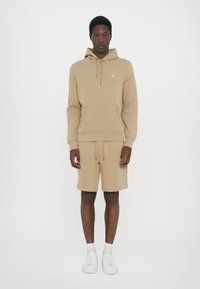 Beige katoenen hoodie met kangoeroezak, bijpassende shorts en witte sneakers. Voorzien van trekkoorden en logo op de borst. Strakke, casual uitstraling.