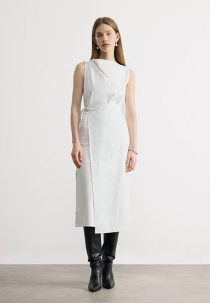 Mladá žena stojí a má na sobě bílý midi šaty bez rukávů s pásem v pase a černé kozačky až po kolena na bílém jednobarevném pozadí.