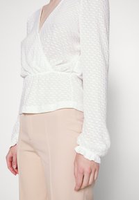 Blusa branca texturizada com decote em V profundo, mangas longas bufantes e cintura franzida, combinada com calças beiges justas.