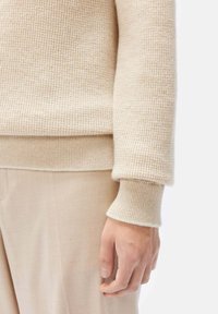 Beige ribbat stickad tröja med långa ärmar och en åtsittande midja, i kombination med ljusbeige byxor, med en närbild på ärmen och handen.