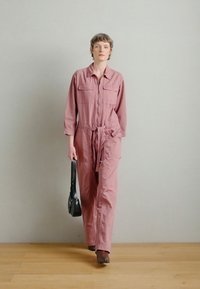 Rózsaszín overál hosszú ujjakkal, cipzáras záródással, két elülső zsebbel és megkötős derékkal. A modell egy kis fekete táskát visel, és fa padlón áll.