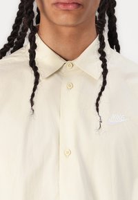 Πουκάμισο με κουμπιά σε ανοιχτό μπεζ ύφασμα; διαθέτει θεσπέδιο γιακά, μία τσέπη με λευκό刺αρισμένο λογότυπο Nike και ορατά κουμπιά.