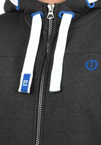 Sudadera gris con cremallera frontal, cordones blancos, acentos azules en la capucha y un pequeño logo azul. Tejido suave con un aspecto texturizado.