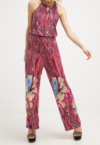 Vrouw in een mouwloze haltertop en wijde broek met rode draaikolk en groot meerkleurig bloemenpatroon, zwarte plateausandalen.