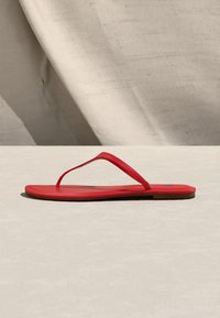Sandale tongs plate rouge avec fines lanières en forme de bride, vue de côté sur une surface neutre avec un fond en tissu drapé.