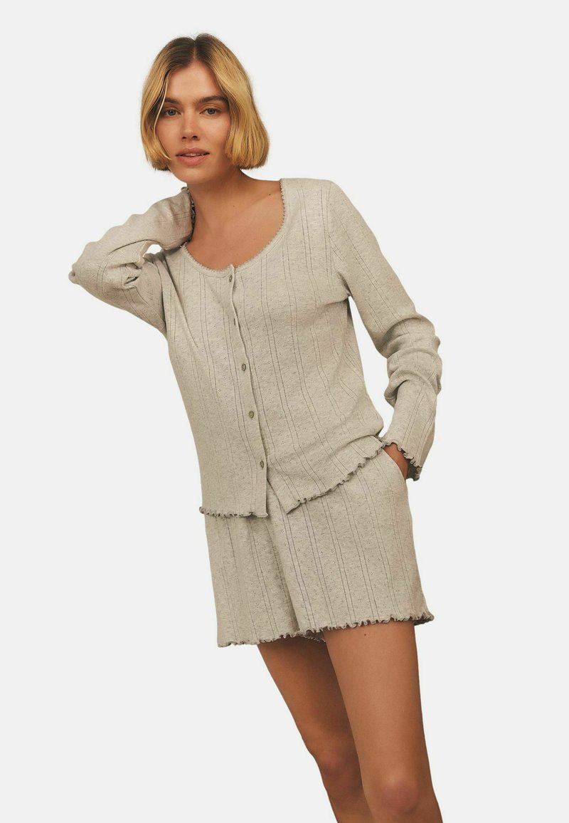 Kvinna med kort blont hår bär en beige räfflad knäppt cardigan och matchande shorts med kantiga kanter, som poserar mot en enfärgad bakgrund.