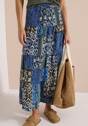 Kobieta nosząca niebieską maxi spódnicę w patchworkowy wzór, beżowe wsuwane buty, trzymająca plecioną słomianą torbę z białymi uchwytami.