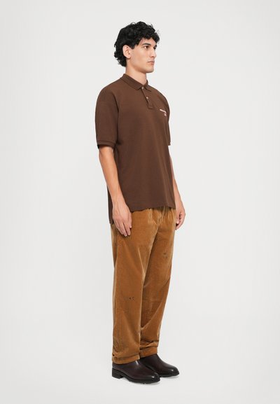 Camisa polo castanha com colarinho, apresentando um logótipo discreto. Combinada com calças de veludo cotelê castanhas claras e botas de tornozelo pretas.