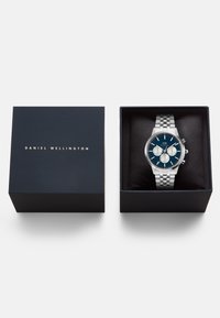 Orologio in acciaio inox argento con quadrante blu, dotato di tre sottoquarzi, contenuto in una scatola nera con testo dorato sopra.