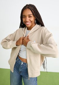 Beige zip-up hette med snøring, frontlommer og ribbede mansjetter, lagd over en grå skjorte, sammen med høytidskjær blå jeans.