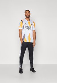 Maglia a righe bianche e gialle con accenti grigi e logo nero, abbinata a pantaloni neri con strisce laterali bianche e scarpe nere.