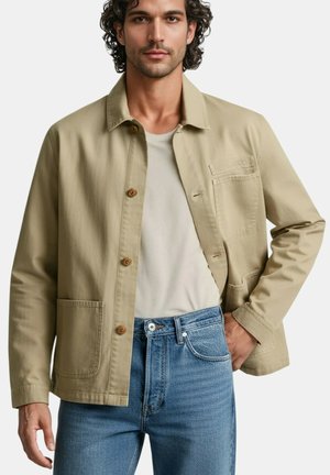 Veste en coton beige avec fermeture à boutons, une poche poitrine et deux poches avant. Portée avec un jean en denim bleu et un t-shirt clair.