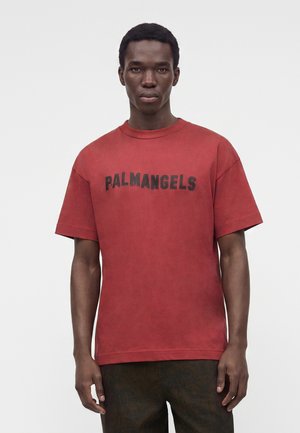 Млад мъж, носещ изтъркан червен T-shirt с "PALMANGELS" в черни букви, стои срещу едноцветен светъл фон.