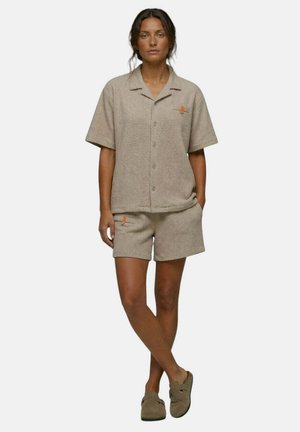 Femme debout, bras détendus, portant une chemise beige texturée à manches courtes avec des boutons, un short assorti et des chaussures sans lacets sur un fond uni.