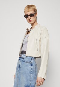 Veste en cuir beige coupée avec col montant, poches zippées sur les côtés et accents côtelés aux épaules, associée à une jupe en denim bleu et un t-shirt graphique.