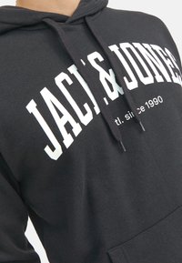 Schwarzer Hoodie aus weichem Baumwollstoff, mit weißer Schrift auf der Brust, die "JACK & JONES" und "est. seit 1990" lautet, sowie mit Kordeln.