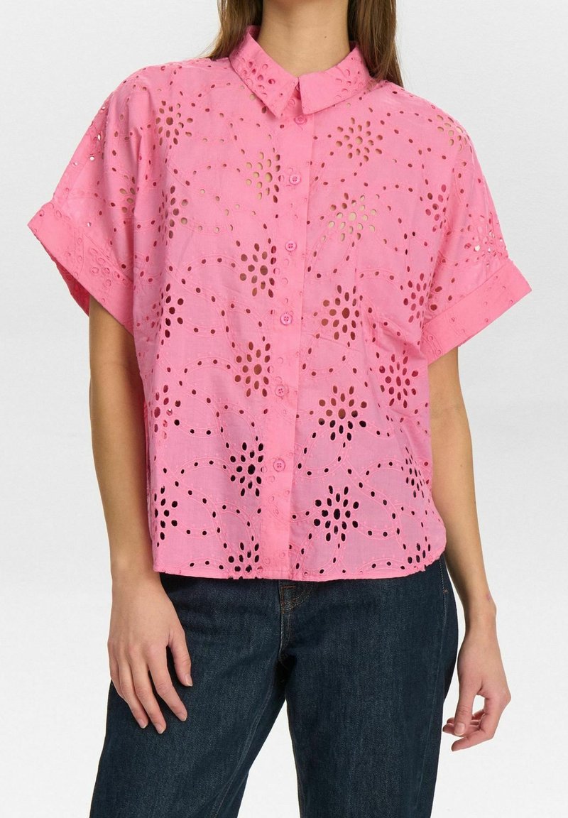 Kobieta ubrana w różową bluzkę na guziki z krótkim rękawem, zdobioną kwiatowymi wycięciami, oraz ciemnoniebieskie dżinsy, stojąca na tle jednolitego tła.